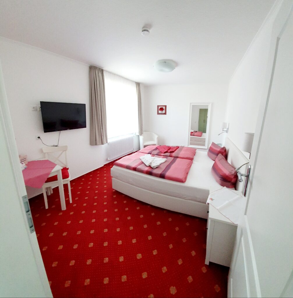 Cozy double room at Haus Hubertus on Borkum with a comfortable bed, red carpet, flat-screen TV, and bright, inviting decor for a relaxing stay.
