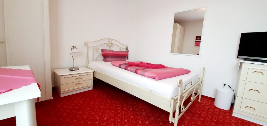 Cozy Einzelzimmer at Haus Hubertus on Borkum, featuring a single bed with pink bedding, bright white furniture, a flat-screen TV, and red carpet.