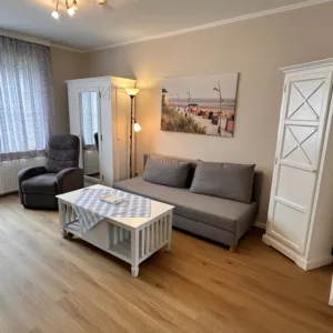 Gemütliches Wohnzimmer im Haus Hubertus auf Borkum mit grauem Sofa, Sessel, weißem Couchtisch und hellem Holzfußboden für einen entspannten Urlaub.