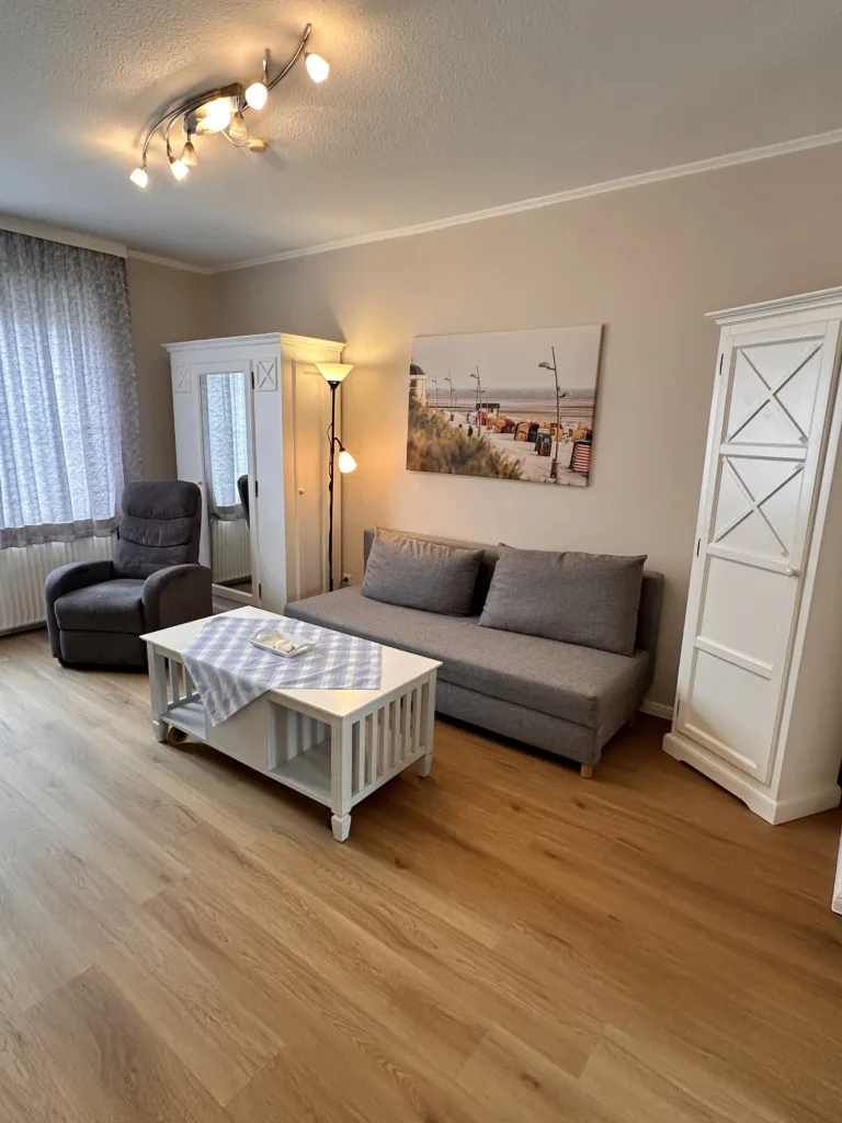 Gemütliches Wohnzimmer im Haus Hubertus auf Borkum mit grauem Sofa, Sessel, weißem Couchtisch und hellem Holzfußboden für einen entspannten Urlaub.