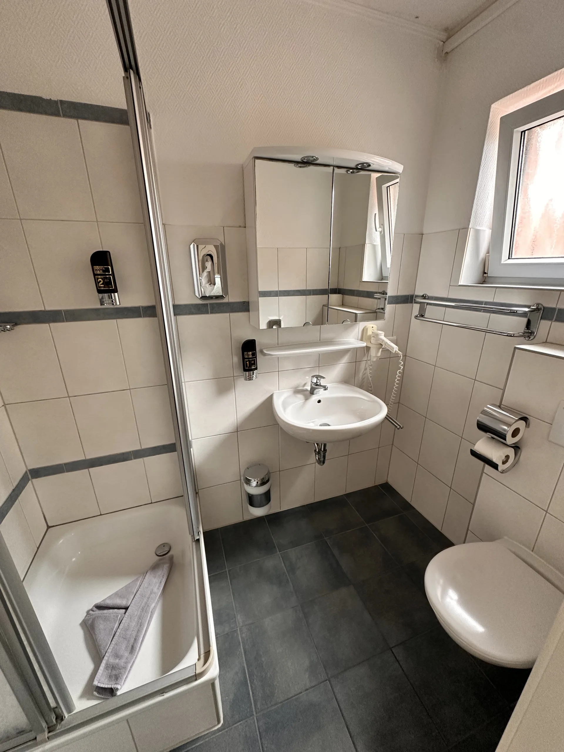 Modern, clean bathroom at Haus Hubertus on Borkum featuring a glass shower, white tiled walls, a sink with mirror cabinet, and dark floor tiles.