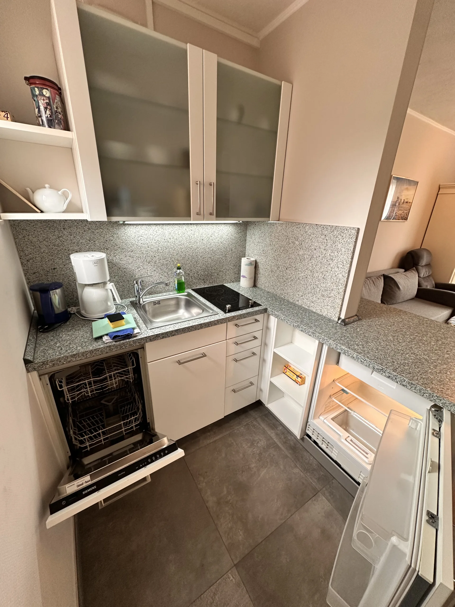 Compact, modern kitchen in a Haus Hubertus holiday flat on Borkum, featuring white cabinets, a dishwasher, fridge, and cozy living area view.