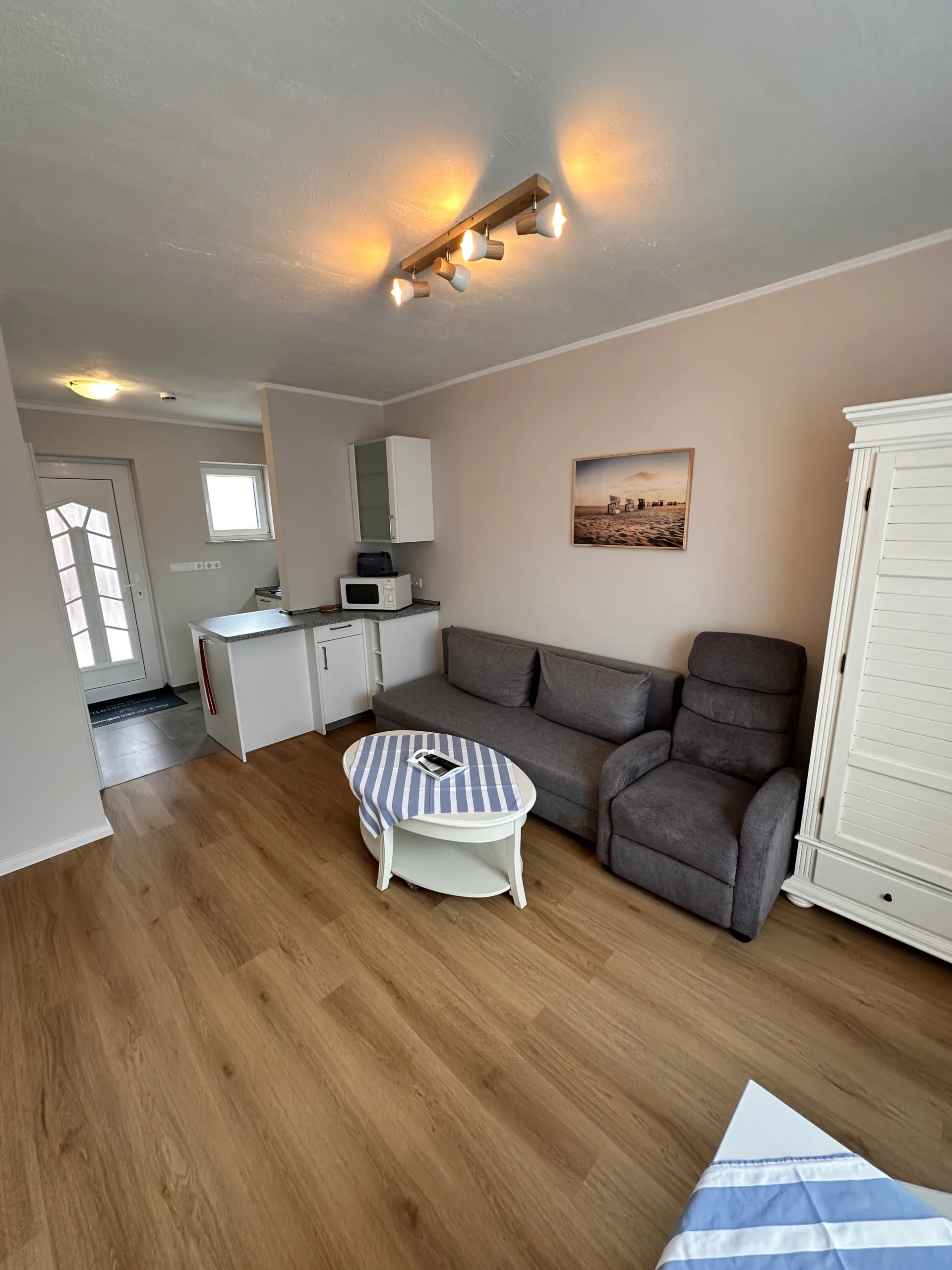 Cozy living area in a Haus Hubertus holiday flat on Borkum, featuring a comfortable grey sofa, armchair, kitchenette, and a beach-themed wall picture.