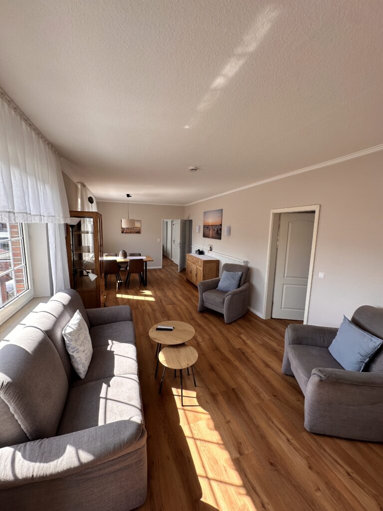 Bright, modern living room in a Hubertus holiday flat on Borkum, featuring comfortable grey sofas, wooden floors, and a cozy dining area.
