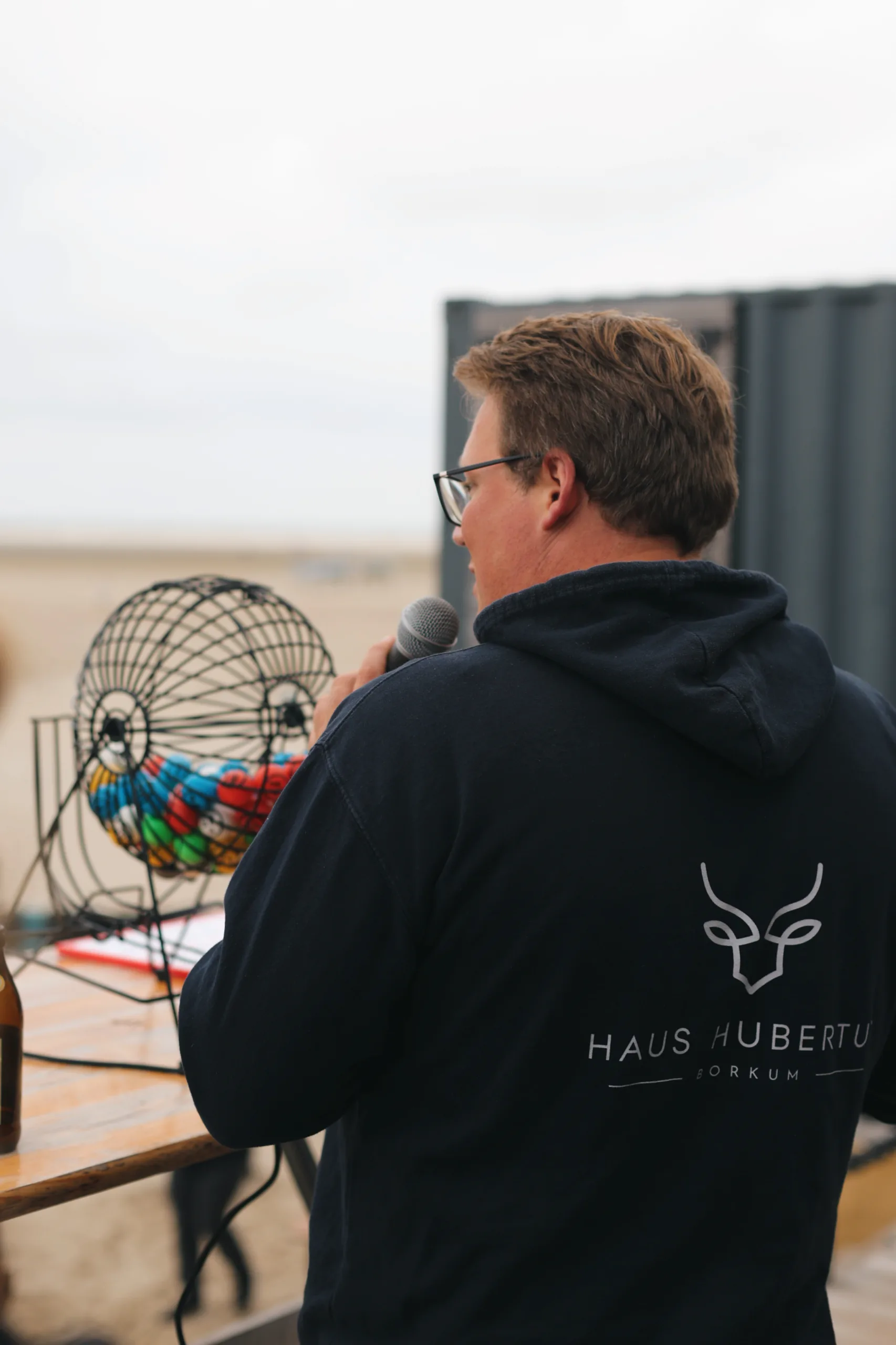 Ein Mann im Haus Hubertus Borkum Hoodie moderiert mit Mikrofon vor einer Lostrommel bei einer Veranstaltung am Strand von Borkum.